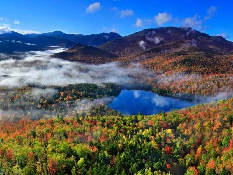 Picture from the top of a mountain of green pine trees amongst fall foliage and in the middle of the picture is a blue lake along the photo is fog laying in the valleys of the mountains