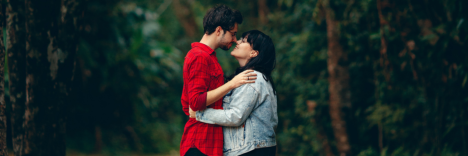 A couple kissing as they stand in front of the pine trees.