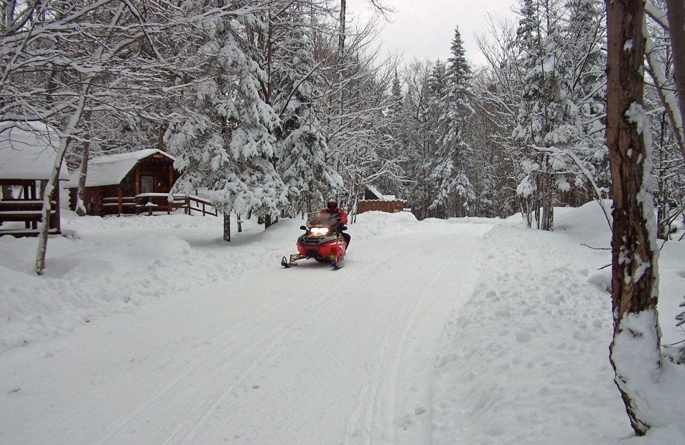 A snowmobiler driving pas wooden cabins on a trail with trees with freshly fallen snow on them around him