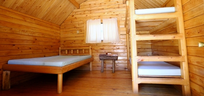 The inside of a cottage with all wood flooring, walls, and ceiling with a window with soft white curtains and a wooden bed next to a night stand and on thee other side is a wooden bunk bed