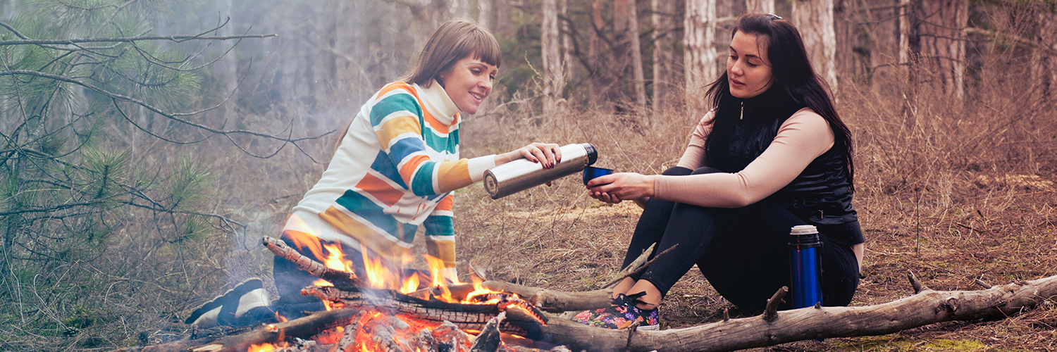 Two women sitting by the campfire having something to drink.