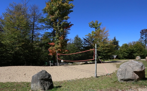 An outdoor volleyball court with trees to ones side a grass to the other with large boulders that you can sit on