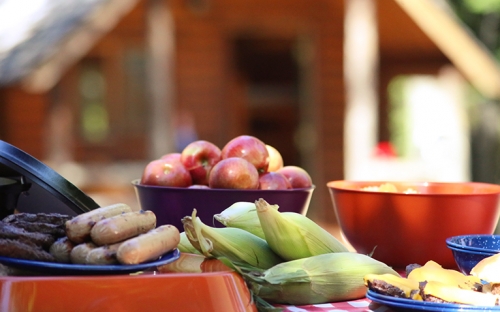 A picnic table with corn on the cob, hotdogs, hamburgers, and a bowl of apples on it with a wood cabin in the background