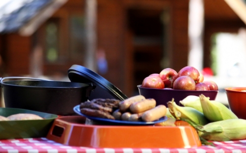 A picnic table with corn on the cob, hotdogs, hamburgers, and a bowl of apples on it with a wood cabin in the background