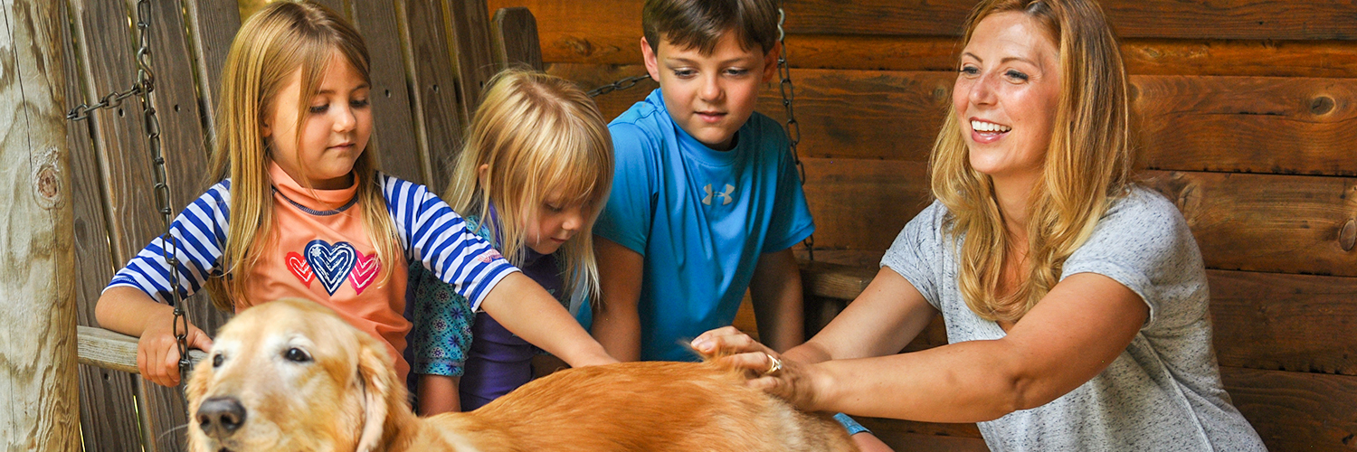 A mother and her kids are sitting on the porch and are petting their dog.