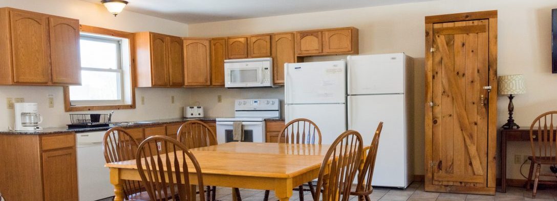 A kitchen with wooden cabinets two refrigerators, a microwave, a dishwasher, a stove, and a coffee maker in the middle of the room is a wooden kitchen table with wooden chairs, against the far wall is a wooden closet and next to that is a desk with a lamp on it and a chair passed into the desk