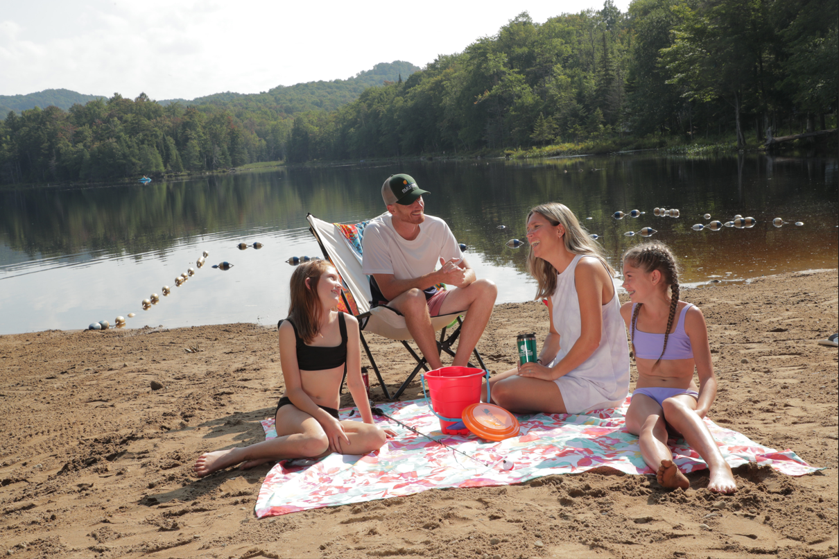 family sitting around a beach towel on a sandy beach with water the lake and green wooded area behind them